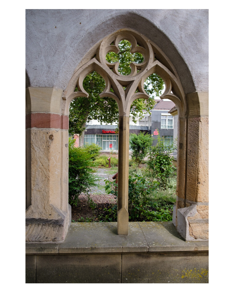 Foto im Hochformat. Ein Fenster in einer steinernen Ruine. Das Fenster läuft oben spitz zu und hat oben verschnörkelte Verzierungen. Durch das Fenster ist eine moderne Kreissparkasse zu sehen. 