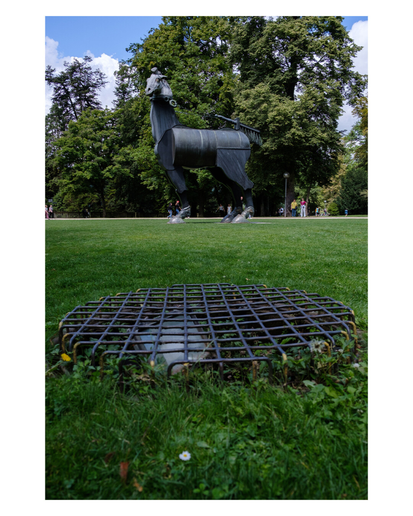 Foto im Hochformat. Eine grüne Wiese, im Vordergrund ist eine runde vergitterte Abdeckung im Boden. Weiter hinten auf der Wiese ist eine große Skulptur aus Metall. Es ist ein Pferd, welches vorne nur ein Bein Hand und statt Schweif hat es einen Kehrbesen. Dahinter sind noch Bäume eines Parks, der Himmel ist leicht bewölkt. 