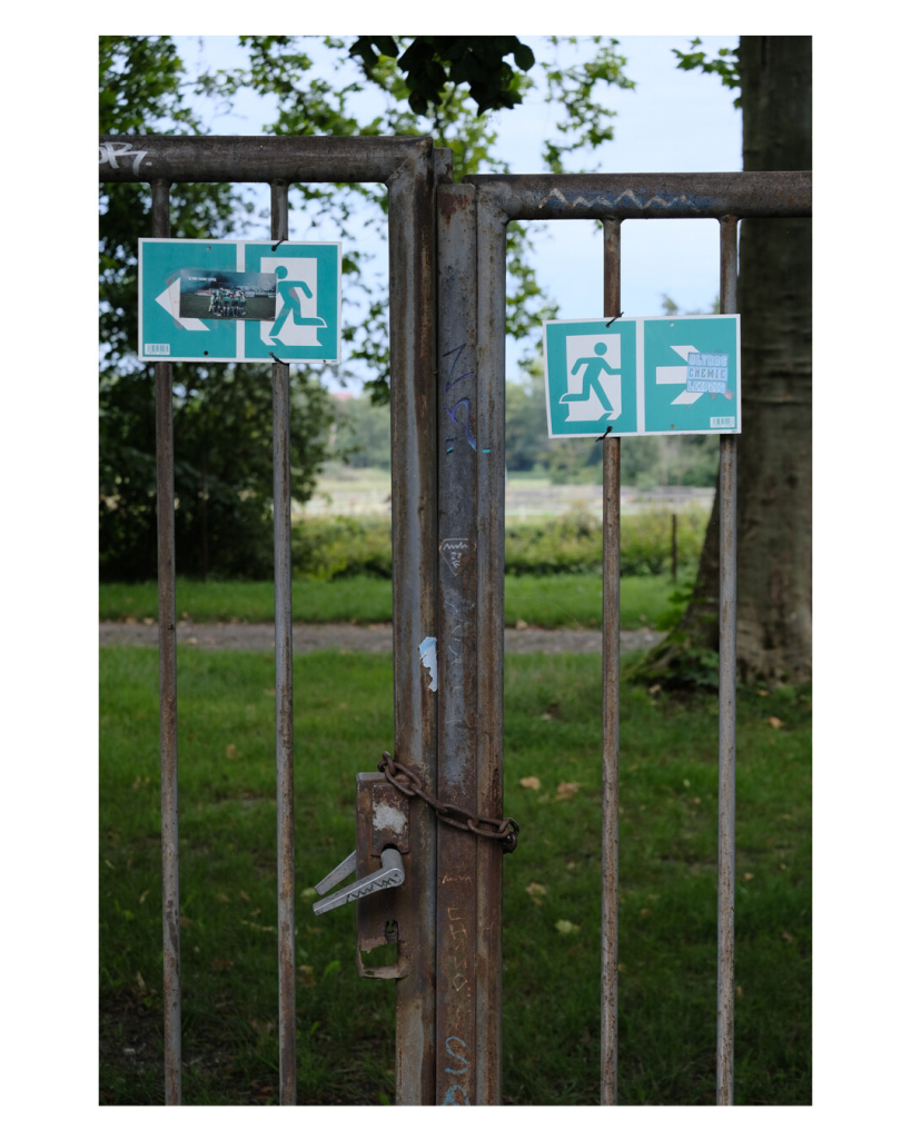 Foto im Hochformat. Ein Gittertor, dahinter ist leicht unscharf ein Park. Das Tor ist mit einer Kette abgeschlossen. Sowohl links als auch rechts hängt ein Notausgangschild, welches in die jeweilige Richtung zeigt. 