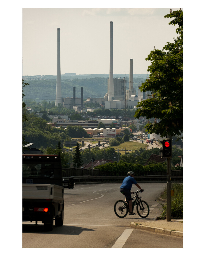 Foto im Hochformat. Teleaufnahme, Blick ins Neckartal auf das Altbacher Kraftwerk mit drei riesigen weißen Schornsteinen. Im Vordergrund steht ein Laster an einer roten Ampel, die von einer Fahrrad fahrenden Person überquert wird. 