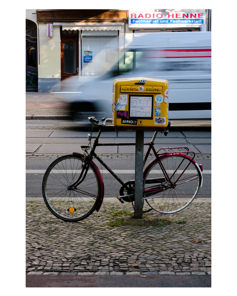 Foto im Hochformat. An einer Straße ist ein gelber Briefkasten auf einem Pfosten. Er ist komplett mit Aufklebern beklebt, auf der Klappe stehe Liebesbriefe. Am Pfosten lehnt ein rotes Rad. Auf der Straße fährt ein weißer Transporter, durch die lange Belichtung ist dieser verwischt, lässt sich aber erkennen. Auf der anderen Straßenseite ist ein Elektronikgeschäft. Es heißt Radio Henne. 