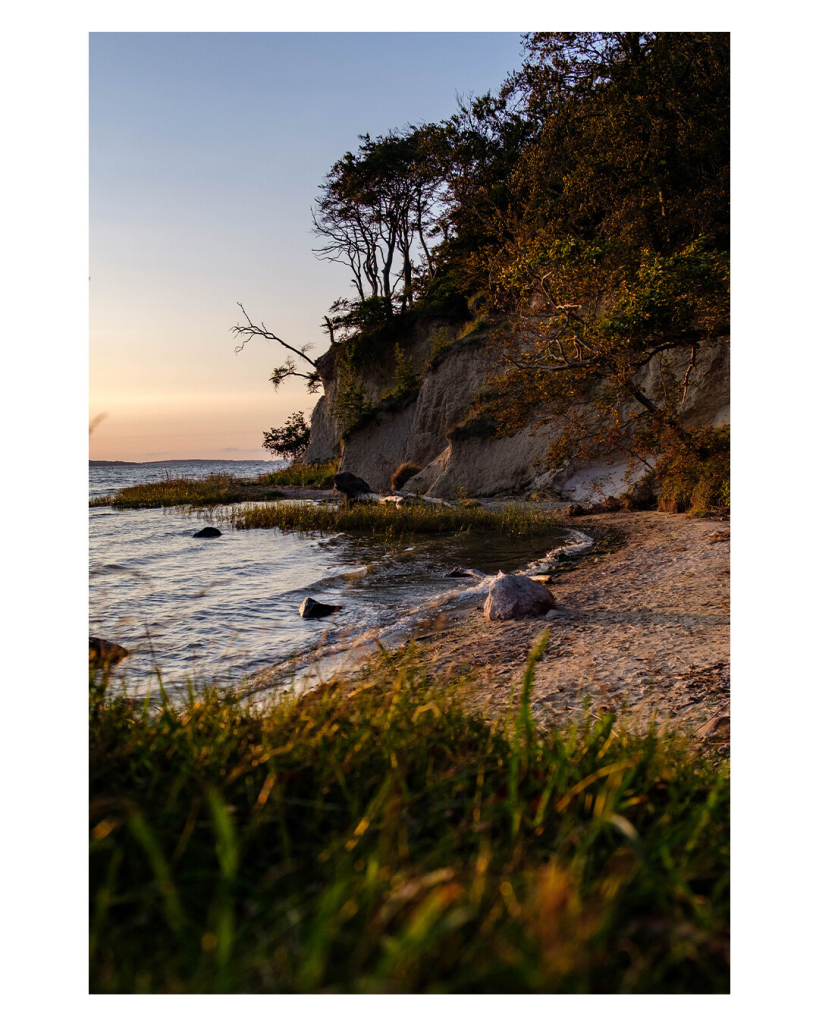 Foto im Hochformat. Blick auf eine kleine Bucht mit Sandstrand am Meer. Am Strand ist kein Mensch. Im Vordergrund ist leicht unscharf ein bisschen Wiese im Bild. Der Himmel beginnt sich schon leicht rötlich zu färben am Horizont, bald ist Sonnenuntergang. 