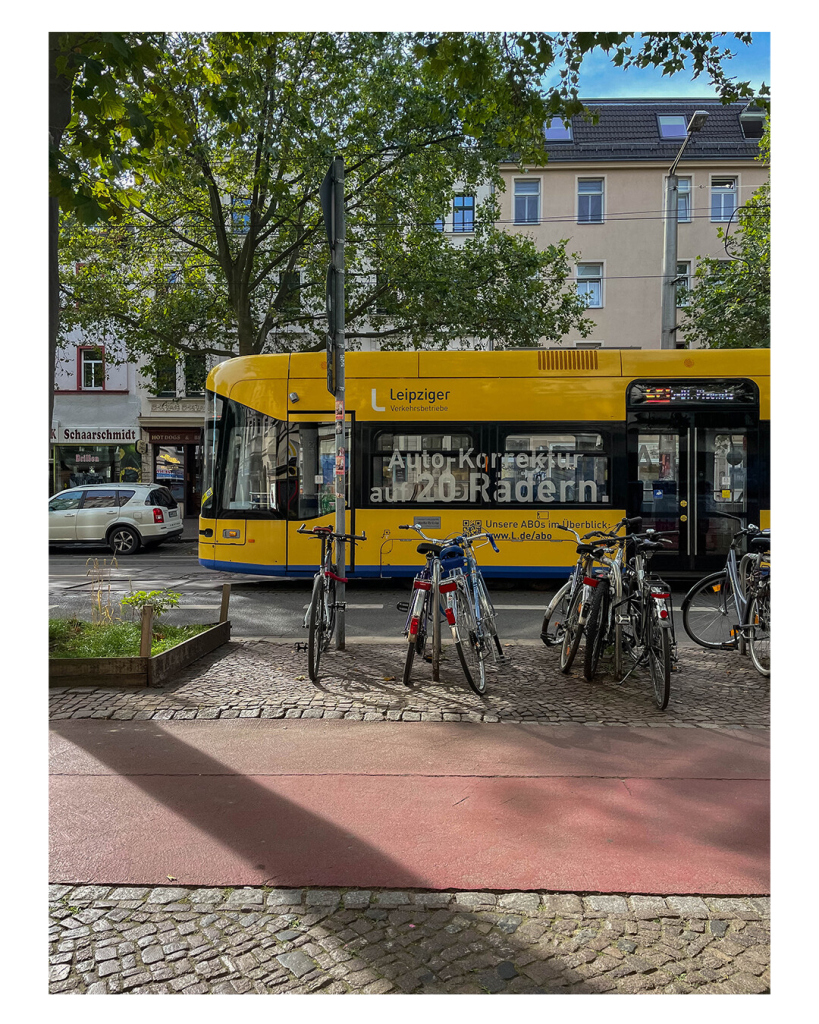 Foto im Hochformat. Eine Straße in Leipzig, eine Straßenbahn fährt von links nach rechts durchs Bild, darauf steht „Auto-Korrektur auf 20 Rädern“. Im Vordergrund ist Geh- und Radweg, an Fahrradständern stehen einige Fahrräder. Es ist Sommer und sonnig. 