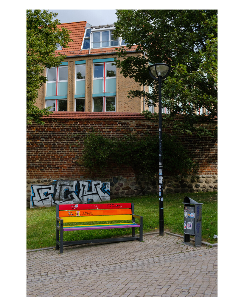 Foto im Hochformat. Ein Parkbank, deren einzelnen Latten und Regenbogenfarben angemalt sind. Daneben eine Straßenlaterne und ein Mülleimer, alles steht vor einer Mauer auf der ein Graffiti ist. Laterne und Mülleimer sind beide mit Unmengen an Aufklebern beklebt. 