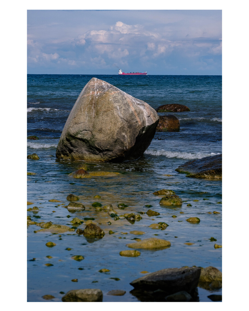 Foto im Hochformat. Blick aufs Meer, im Vordergrund ist Watt, bedeckt mit vereinzelten Steinen. Etwas weiter hinaus ist ein großer Felsen. Am Horizont auf dem Meer fährt ein großes Containerschiff. Das Meer am Horizont ist nur ein bisschen über der Spitze des Felsen, es macht also ein bisschen den Eindruck, als ob das Schiff gleich gegen den Felsen fährt. 
Der Himmel ist leicht bewölkt. 