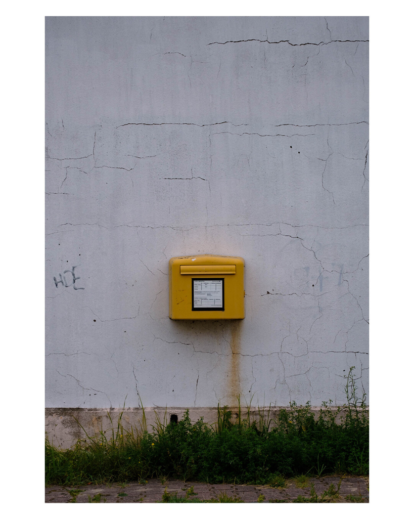 Foto im Hochformat. Eine weiße brüchige Hauswand, daran ist ein einzelner öffentlicher gelber Briefkasten montiert. Auf der rechten Seite geht eine gelb-bräunliche Spur vom Briefkasten Richtung Boden. Zwischen Boden und Hauswand wächst Gras und Unkraut. Links an der Wand steht HDE, lieblos hingeschrieben. 