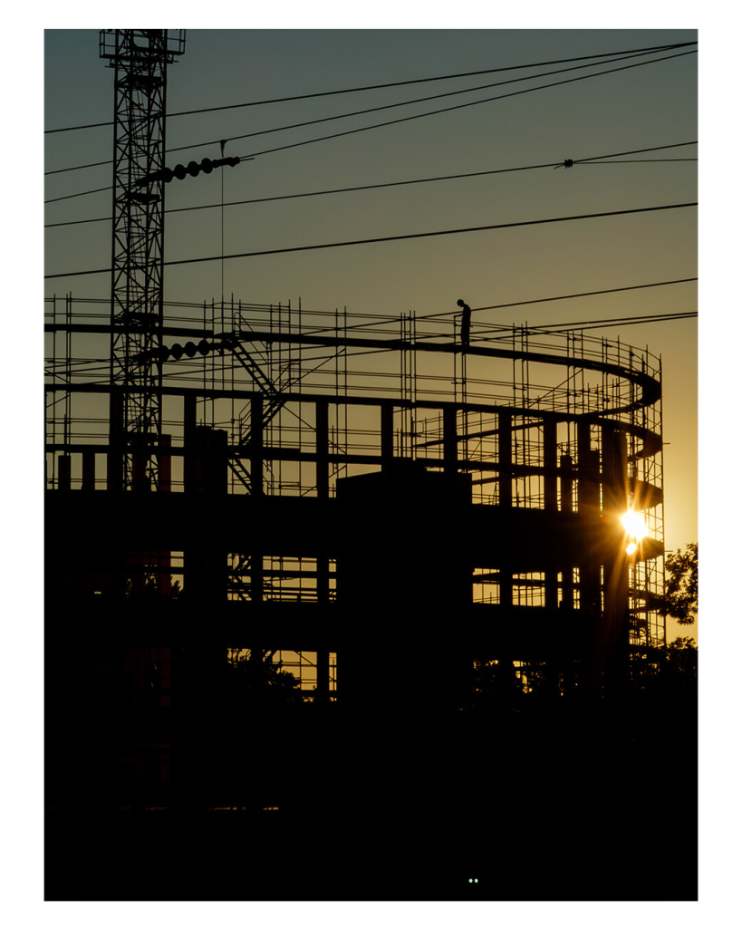 Foto im Hochformat. Baustelle, ein rundes Gebäude wird errichtet. Das nackte Skelett des Gebäudes steht vor der untergehenden Sonne. Es ist nur eine schwarze Silhouette vor dem blau-gelben Himmel. Die Sonne ist knapp über dem Horizont und zur Hälfte vom Gebäude verdeckt. Ganz oben auf dem Gerüst des Gebäudes ist die Silhouette einer Person mit Blauhelm, die nach unten schaut. Der untere Teil des Bildes ist nur schwarzer Schatten. 