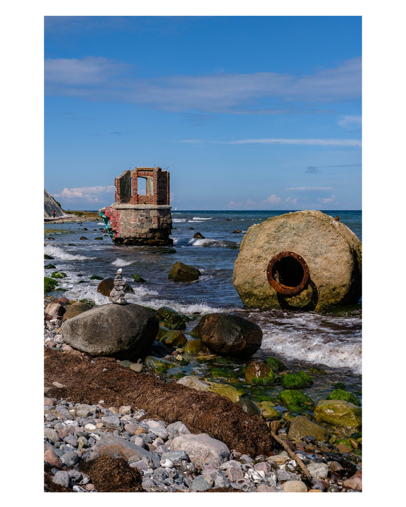 Foto im Hochformat. Ein steiniger Strand am Meer. Auf einem Felsen ist ein kleiner Turm aus aufeinandergestapelten Stein. Im Meer steht ein großes Steinrad mit einem rostig metallenen Loch in der Mitte. Weiter hinten im Meer ist ein kleines kaputtes Haus, welches auf einer Plattform steht. 
Der Himmel ist leicht bewölkt. 