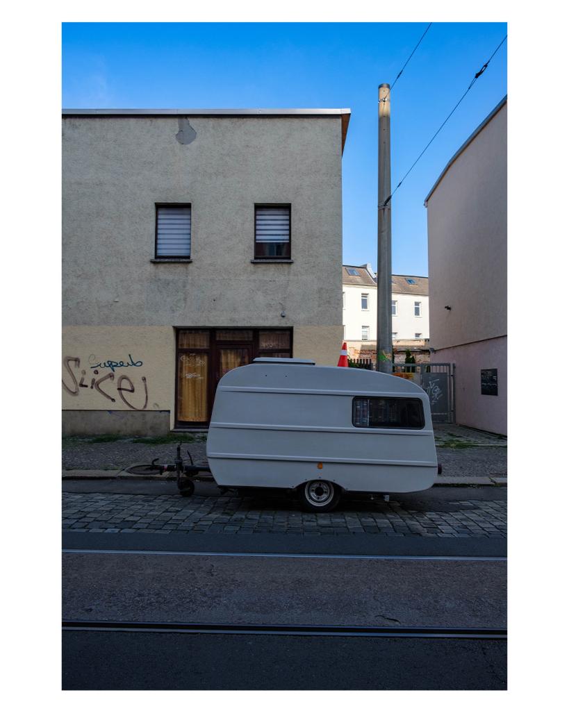 Foto im Hochformat. An einer Straße steht ein kleiner weißer Wohnwagen. Auf dessen Dach platziert ist ein orange-weißer Warnkegel. Hinter dem Wohnwagen ist ein grau-gelbliches altes Gebäude auf dessen Wände mehrere Worte gesprüht sind. Der Himmel ist strahlend blau. 