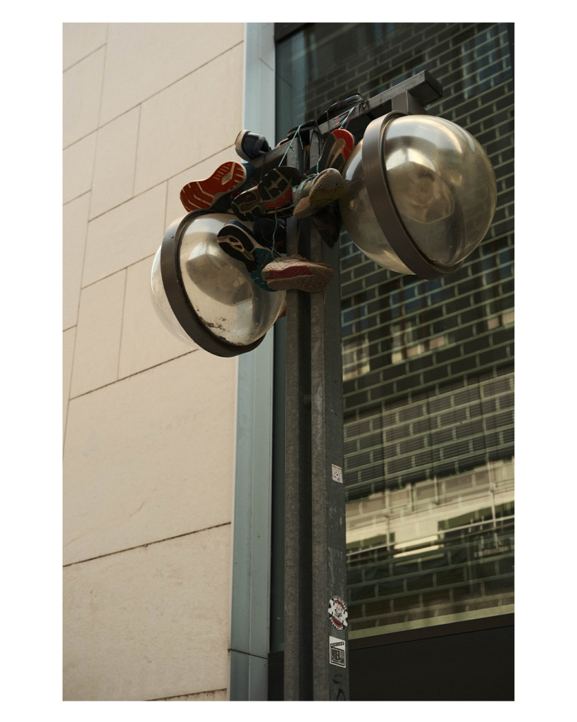 Foto im Hochformat. Eine Straßenlaterne mit links und rechts jeweils einer Glaskugel in der die Lampe ist. In der Mitte von beiden, über den Pfosten der Laterne, sind mehrere zusammengeknotete Schuhpaare gehängt. 