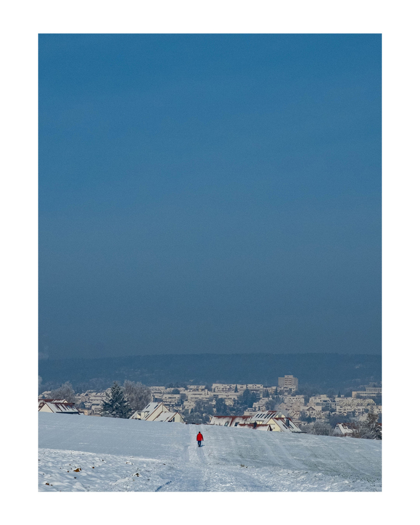 Foto im Hochformat. Ein mit Schnee bedecktes Feld. Am Horizont sind die Dächer einer Stadt, dahinter erhebt sich Wald, darüber ist der klare blaue Himmel, der den Großteil des Bildes einnimmt. Mittig auf den Feldern ist ein zugeschneiter Weg. Darauf läuft eine einzelne Person, die sich mit ihrer roten Jacke deutlich vom Schnee abhebt. 