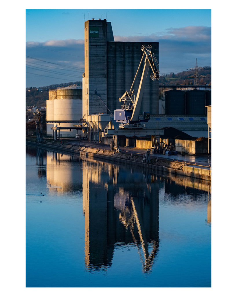 Foto im Hochformat. Ein Hafenbecken. Am Ufer steht ein großes graues Industriegebäude, davor sind Lagerhallen und ein großer Kran. Alles spiegelt sich im leicht gekräuselten Wasser auf dem auch ein paar Enten schwimmen. 