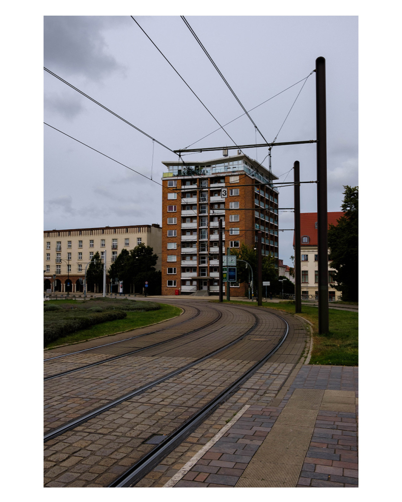 Foto im Hochformat. Straßenbahnschienen gehen von links unten im Bild in einer Kurve link aus dem Bild. Darüber verlaufen parallel dazu Leitungen. Im der Kurve der Schienen steht eine großes altes Wohngebäude aus rotem Backstein mit vielen Balkons. Der Himmel ist bewölkt. 