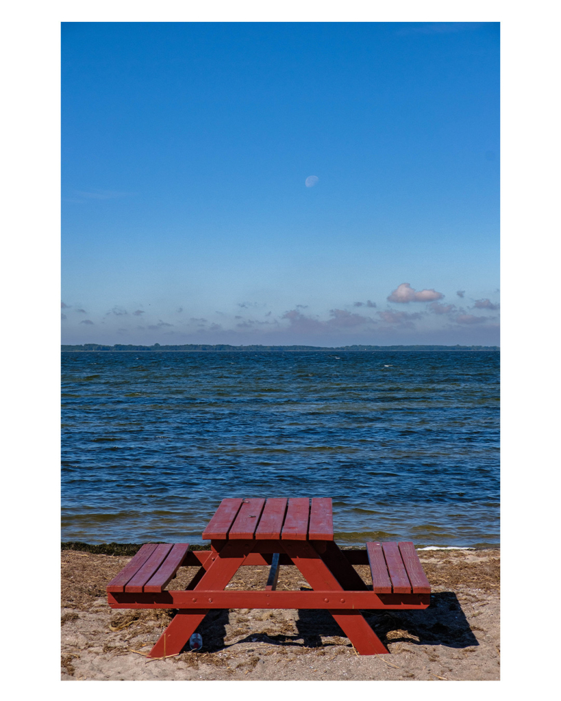 Foto im Hochformat. Blick aufs Meer, im unteren Teil des Bildes ist Strand, auf dem eine rote Picknick-Bank steht. In der Mitte ein Tisch, rechts und links eine Bank. Dahinter ist das Meer, der Horizont ist genau in der Mitte des Bildes. Über dem Horizont sind ein paar Wolken, ansonsten ist der Himmel blau. Mitten am Himmel ist der Mond auszumachen. 