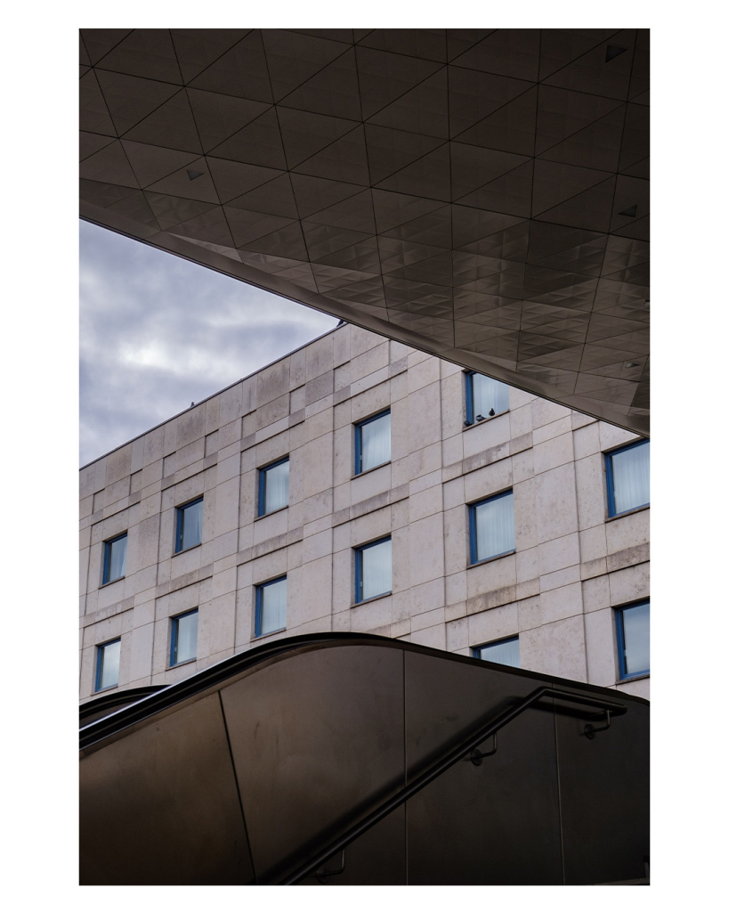 Foto im Hochformat. Blick auf ein Gebäude von einer Unterführung aus. Das Gebäude ist ein grauer Betonklotz mit Reihen von gleichen Fenstern. Der obere Teil des Bildes ist eine Decke, der untere Teil ist eine Wand, an der vorne ein Geländer ist und dahinter eine Rolltreppe. Die Decke, das Gebäude und der linke Rand des Bildes bilden ein fast gleichseitiges Dreieck in dem der bewölkte Himmel ist. Die Farben des Fotos sind bedeckt gehalten, grau und metallisches blau. 