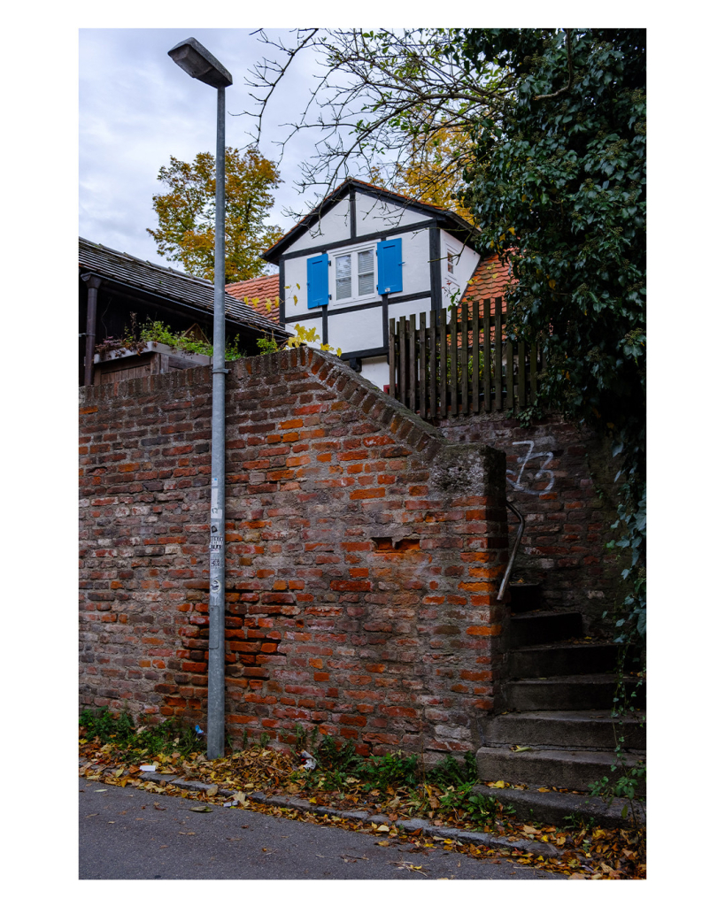 Foto im Hochformat. Eine Backsteinmauer, davor steht eine Straßenlaterne, dahinter geht eine Treppe hoch. Oben, über der Mauer schaut der obere Teil eines Fachwerkhauses hervor. Weiß mit braunen Balken. Am einzgen Fenster sind blaue Fensterläden. Es ist Herbst. 