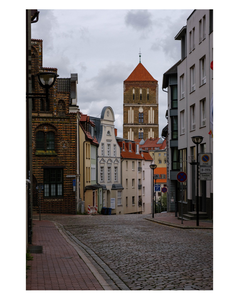 Foto im Hochformat. Eine komplett leere Straße mit Kopfsteinpflaster. Sie geht ein Stück geradeaus, dann einen Berg herunter. Am linken und rechten Straßenrand ist eine Mischung aus alten und neuen Gebäuden. Hinter den Gebäuden ist ein hoher breiter Kirchturm mit spitzem Dach auf dem ein Kreuz ist. Der Himmel ist wolkenverhangen. 