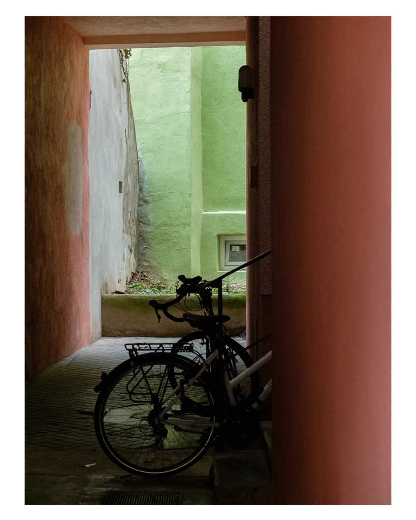 Foto im Hochformat. Blick durch einen Durchgang in einen Hinterhof. Wände und Decke sind in einem altrosa. Am Ende des Durchganges fällt von oben Sonne herab, die Wände sind leuchtend hellgrün. Im Durchgang stehen Fahrräder. 