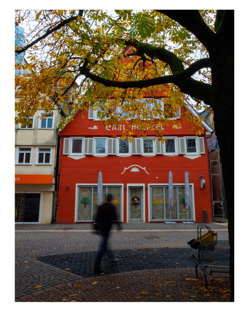 Fotobücher Hochformat. Ein rot verputztes Gebäude mit weißen Fenstern. Davor ist eine Fußgängerzone. Das Gebäude wird von einem Baum und dessen Ästen eingerahmt. Die noch wenigen Blätter am Baum sind herbstlich gelb und grün. Eine Person läuft durchs Bild. Durch längere Belichtungszeit ist diese leicht verschwommen. 