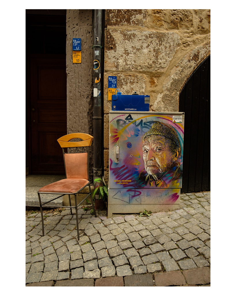 Foto im Hochformat. Eine Kopfsteinpflasterstraße an einem alten Haus. Vor einem Hauseingang steht ein mit Leder bezogener Stuhl mit Holzlehne.  Dahinter ist eine Regenrinne. Daneben ist ein Strommasten auf dem ein buntes Portrait von Michel Tournier (1924 - 2016) gemalt wurde. Auf dem Strommasten steht ein blauer Pappkarton auf dem Mozzarella steht. 