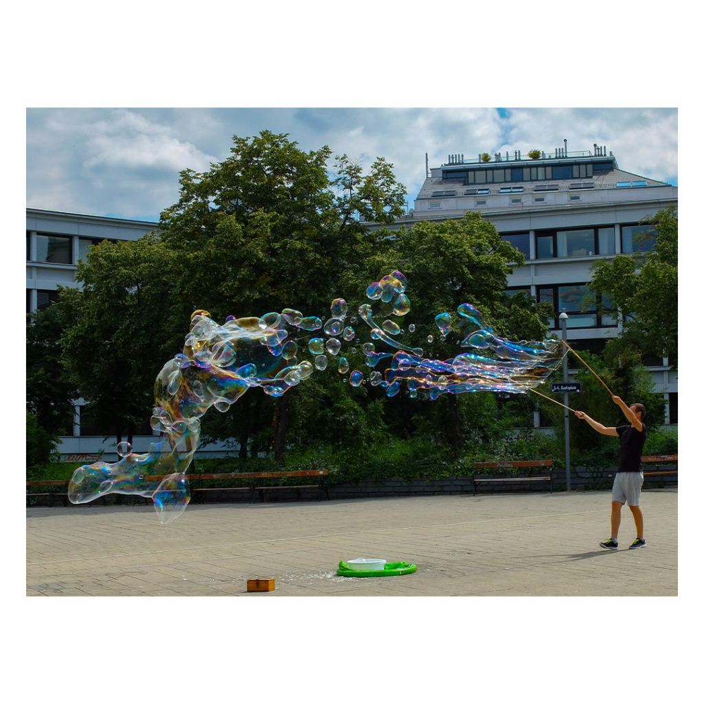 Foto im Querformat. Ein*e Straßenkünstler*in macht mit zwei Stöcken und Schnüren riesige Seifenblasen bzw. viele kleinere Seifenblasen, die sich über mehrere Meter erstrecken. 