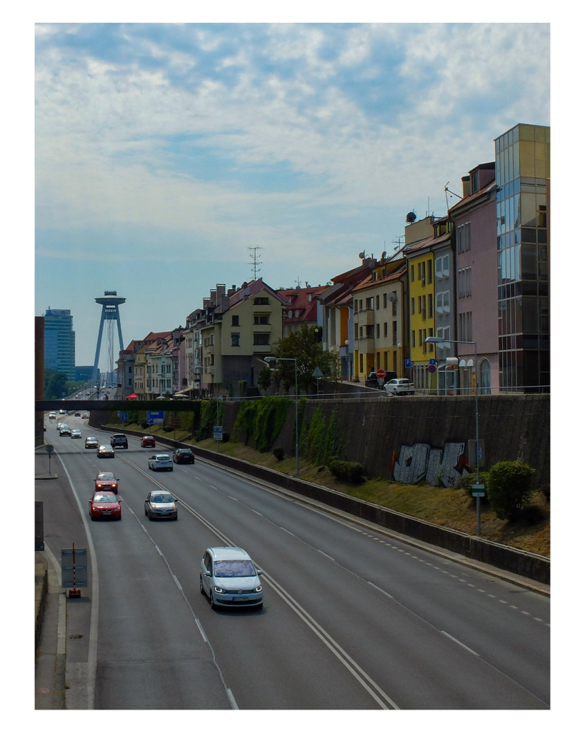 Foto im Hochformat. Eine vierspurige befahrene Straße. Sie führt Richtung Horizont, wo ein großer Turm steht. Der Turm ist auf zwei großen Pfeilern und sieht aus wie ein UFO. Auf der rechten Seite der Straße sind hohe Wohnhäuser. Der Himmel ist bewölkt. 