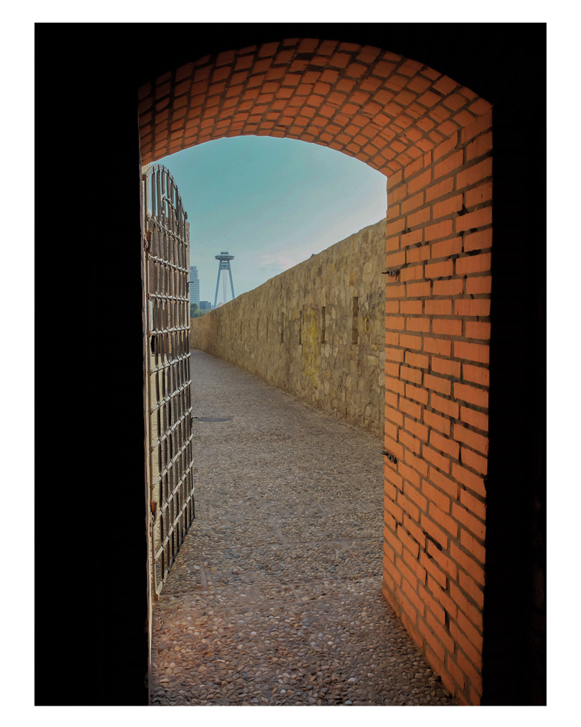 Foto im Hochformat. Blick aus einer Tür in einer dicken Backsteinmauer. Der Türrahmen ist oben rund, die Türe selbst ist eine Gittertür, die offen steht. Dahinter ist rechts eine hohe Mauer. Darüber ist am Horizont ein hoher Turm zu sehen, der wie ein Ufo aussieht. Das UFO wird eingerahmt von der Mauer, dem oberen Türrahmen und der Gittertür. Der Himmel ist blau. 