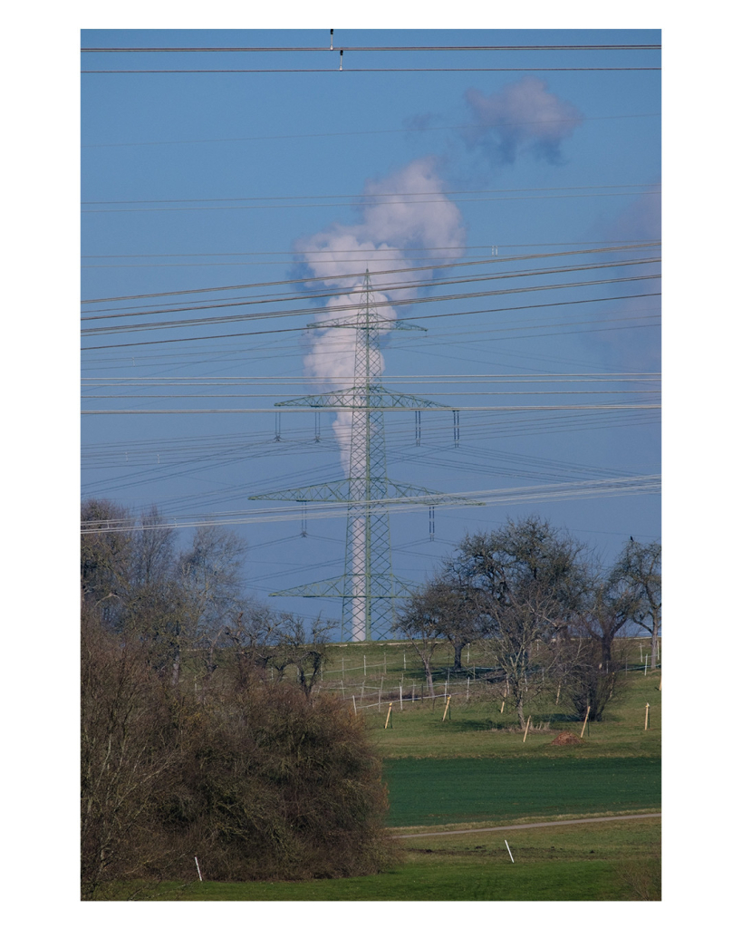 Foto im Hochformat. Eine Wiese, auf der viele Zäune und einige Bäume stehen. Sie geht leicht bergauf. Am Horizont ist ein Hochspannungsmasts. Direkt hinter dem Masten ist ein riesiger weißer Schornstein aus dem dicken Wolken empor steigen. Im Vordergrund und auch sonst im Bild verlaufen Unmengen an Stromleitungen. 