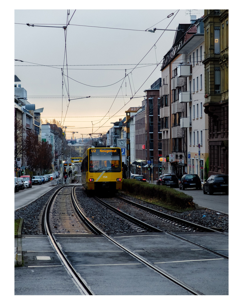 Foto im Hochformat. Straßenbahnschienen verlaufen von vorne nach hinten durchs Bild. Auf der rechten Seite ist eine gelbe U-Bahn mit der gelben Aufschrift U2 Neugereut. Rechts und links ist jeweils eine Straße und daneben jeweils Altbauten. Der Himmel ist bedeckt, davor sind die Oberleitungen der Bahn. Das ganze Bild ist farblich sehr bedeckt, nur die gelbe Bahn sticht heraus. 