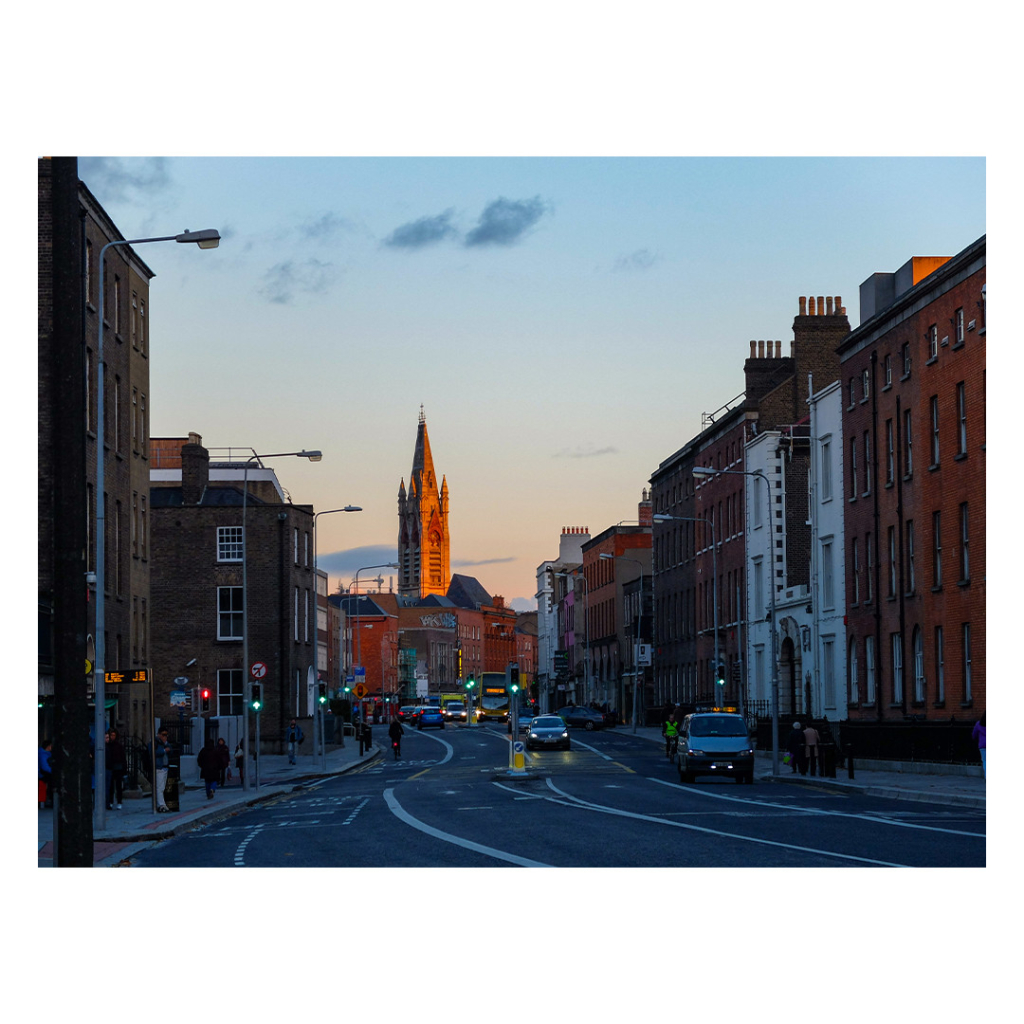 Foto im Querformat. Eine Straße in Dublin, die Straße und die Häuser sind im Schatten. Am Ende der Straße ist ein Kirchturm, dieser wird von der Abendsonne beschienen und leuchtet entsprechend rötlich. Der Himmel ist leicht bewölkt. 