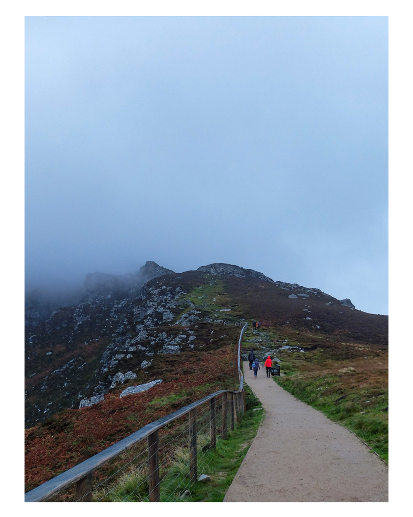 Foto im Hochformat. Die untere Hälfte des Bildes ist ein Berg mit Felsen, teils bewachsen mit Gras. Ein asphaltierter Weg führt nach oben, auf der linken Seite ist ein Geländer. Weiter vorne im Bild laufen Menschen, eine Person davon hat eine rote Jacke an, die heraus sticht. Die obere Hälfte des Bildes sind Wolken, die die Spitze des Berges einhüllen. 