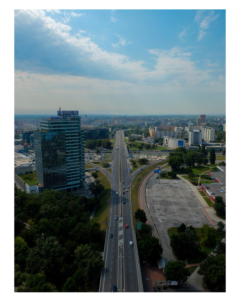 Foto im Hochformat. Blick von oben auf eine Großstadt. Mittig im Bild verläuft eine vierspurige Autobahn Richtung Horizont. Links ist ein großes Hochhaus, am Horizont sind viele Hochhäuser. Der Himmel ist blau, mit einer großen weißen Wolke. 