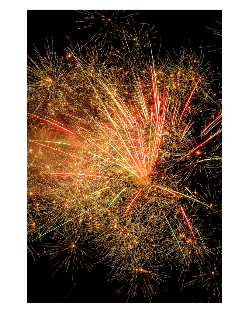 Foto im Hochformat. 
Abgefeuertes Feuerwerk am schwarzen Nachthimmel. Viele dünne lange und kurze Streifen, rot, gelb, grün. 