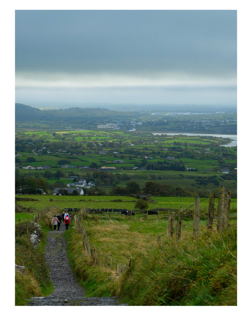 Foto im Hochformat. Blick in ein grünes Tal in Irland. Links unten führ ein Kiesweg nach unten, auf diesem laufen mehrere Personen. Im Tal selbst sind Wiesen, Büsche, Häuser, ein See, am Horizont sind Hügel. Der Himmel ist bewölkt. 