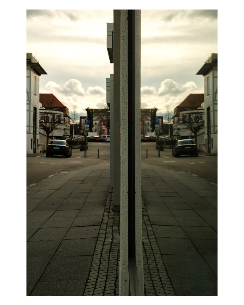 Foto im Hochformat. Eine Straße in einer Stadt, die sich in einem Schaufenster, welches mittig im Bild ist, spiegelt. 