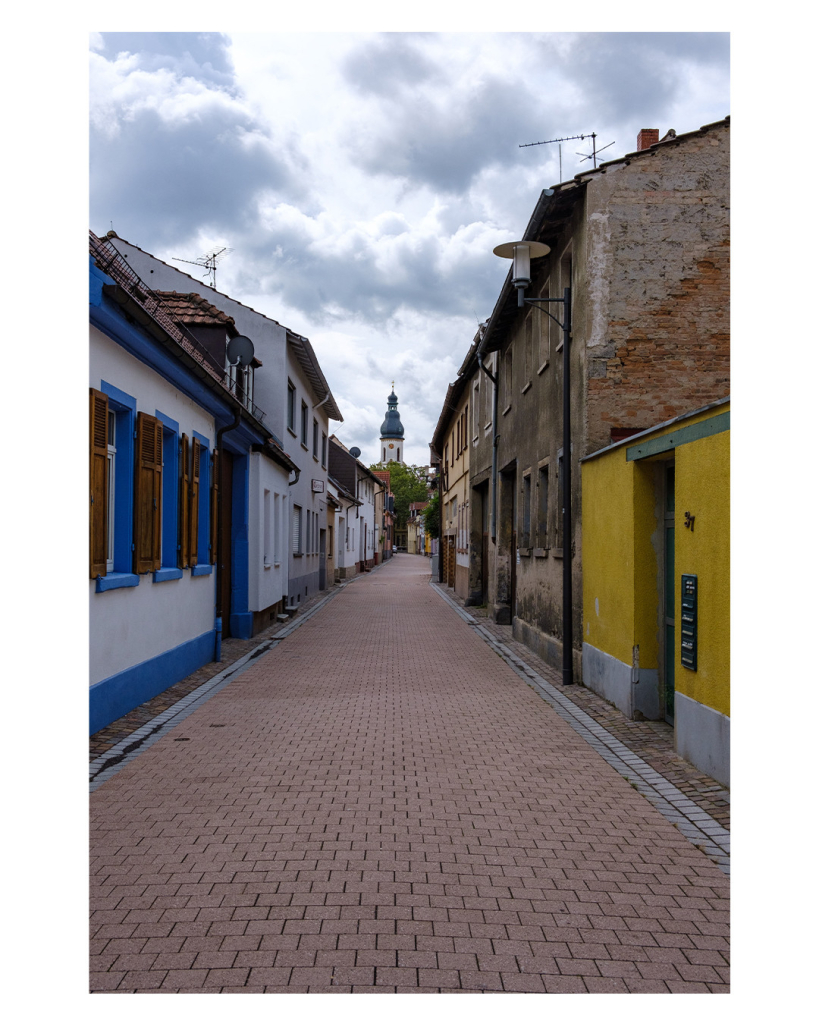 Foto im Hochformat. Blick entlang einer leeren Straße aus rötlichen Steinen. Links und rechts sind Einfamilienhäuser, unterschiedlichen Alters, Bauweise und Farbe. Tendenziell ist alles eher alt, teils Fachwerk. Am Ende der Straße ist ein Kirchturm mit zwiebelförmiger Spitze. Der Himmel ist bewölkt. 