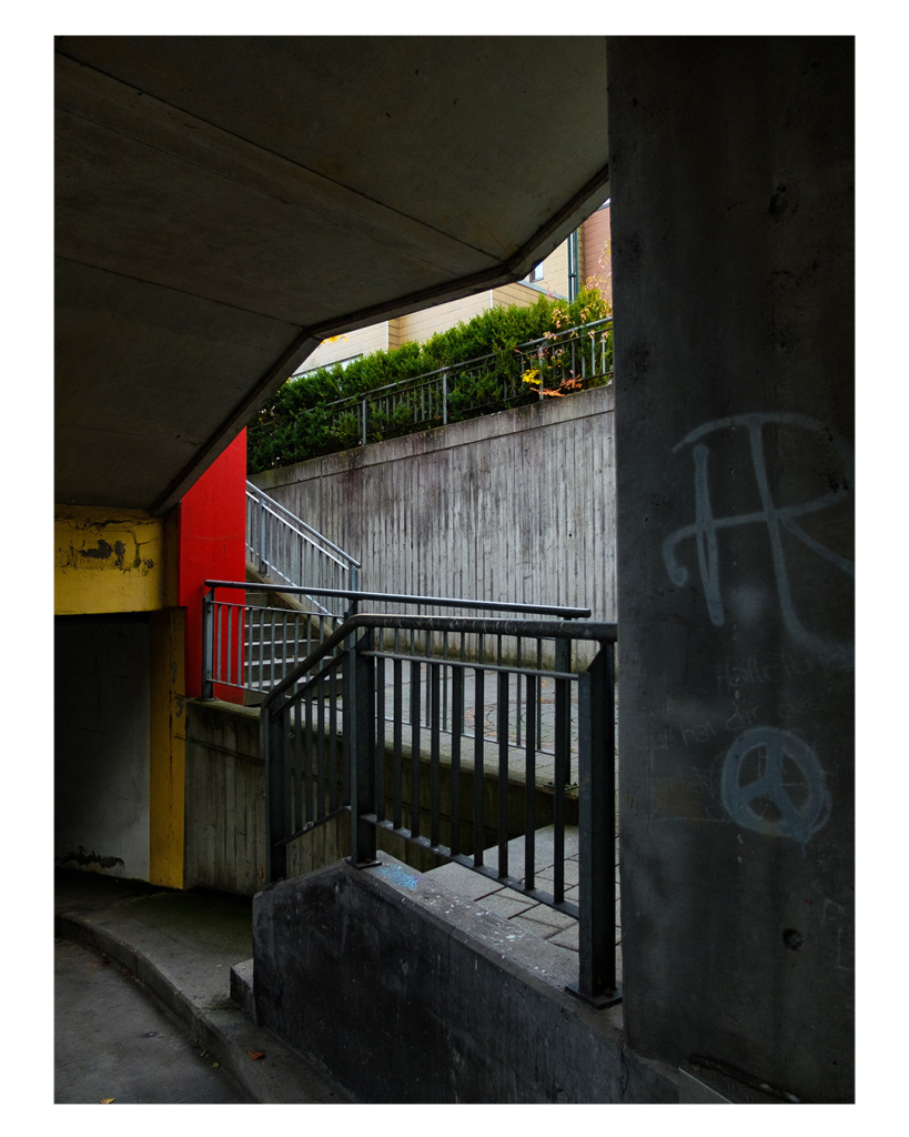 Foto im Hochformat. Blick aus einer Unterführung. Rechts ist eine dicke graue Säule mit schlechten weißen Graffiti. Daneben geht eine kurze r Tee rope runter, dahinter eine Treppe hoch. Alle Wände sind grau bis auf zwei Stellen die gelb und rot sind. 