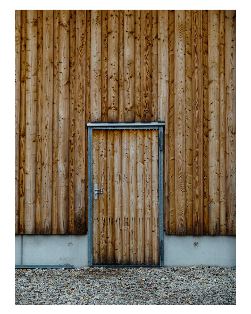 Foto im Hochformat. Eine Wand aus braunen Brettern, die vertikal verlaufen. In der Wand ist eine Türe, ebenfalls mit Brettern, allerdings sind diese etwas dünner. Die Türe hat einen Rahmen aus Metall. Der untere Teil der Wand ist hellgrauer Beton. Vor der Wand ist Kies. 
Alles ist frontal fotografiert, die Wand ist parallel zur Kamera, die Türe ist genau mittig. 