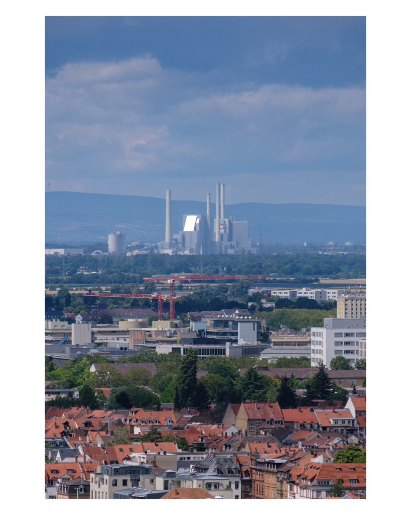 Foto im Hochformat. Eine Teleaufnahme vom Heidelberger Schloss aus. Unten ist die Altstadt im Bild, dahinter dann Gewerbegebiete mit großen Gebäuden, dahinter dann Wälder und dahinter sieht man schließlich das Großkraftwerk Mannheim. Dieses besteht aus riesigen Gebäuden und vier hohen Schornsteinen. Der Himmel darüber ist bewölkt. 