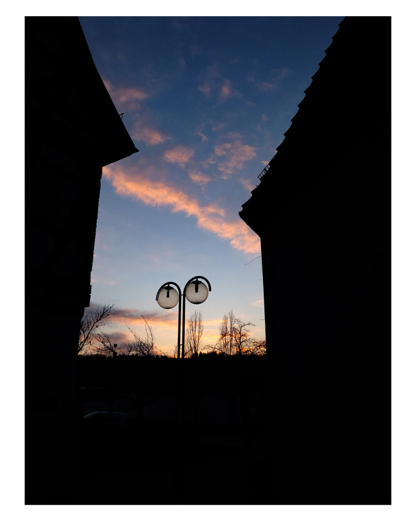 Foto im Hochformat. Abends, die Sonne ist kurz vorm untergehen. Links und rechts ist jeweils ein Haus mit Spitzdach. Unten ist eine Mauer. Alles davon ist nur schwarz im Schatten. Dahinter ist der Abendhimmel, der durch die Hauser und die Mauer die Form des Buchstabens Z hat. In der Mitte steht noch eine Straßenlaterne mit zwei Glaskugeln. Darüber ist ein Streifen Wolken am Himmel. 