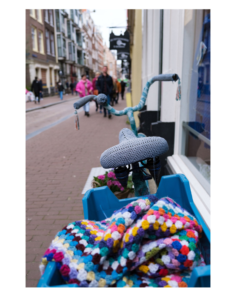 Foto im Hochformat. Eine Straße mit Geschäften. Direkt vor der Kamera steht ein Fahrrad, man sieht von hinten nach vorne über den Lenker die Straße. Das Fahrrad ist komplett überzogen mit gestrickter/gehäkelter Wolle in unterschiedlichen Farben. Auf dem Gepäckträger ist eine blaue Plastikkiste, in dieser liegt eine gehäkelte/gestrickte Decke. Die Schärfe im Bild liegt auf dem Fahrrad, die Menschen in der Straße sind unscharf und nicht zu erkennen. 