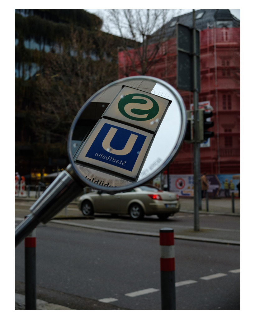 Foto im Hochformat. Der Seitenspiegel eines Motorrollers. Darin spiegelt sich ein S-Bahn und ein U-Bahn-Schild. Ein weißes großes S in einem grünen Kreis und ein weißes großes U in einem blauen Quadrat. Im Hintergrund ist leicht unscharf eine Straße und Häuser in einer Großstadt. Direkt unter dem Spiegel fährt gerade eine Auto entlang. 