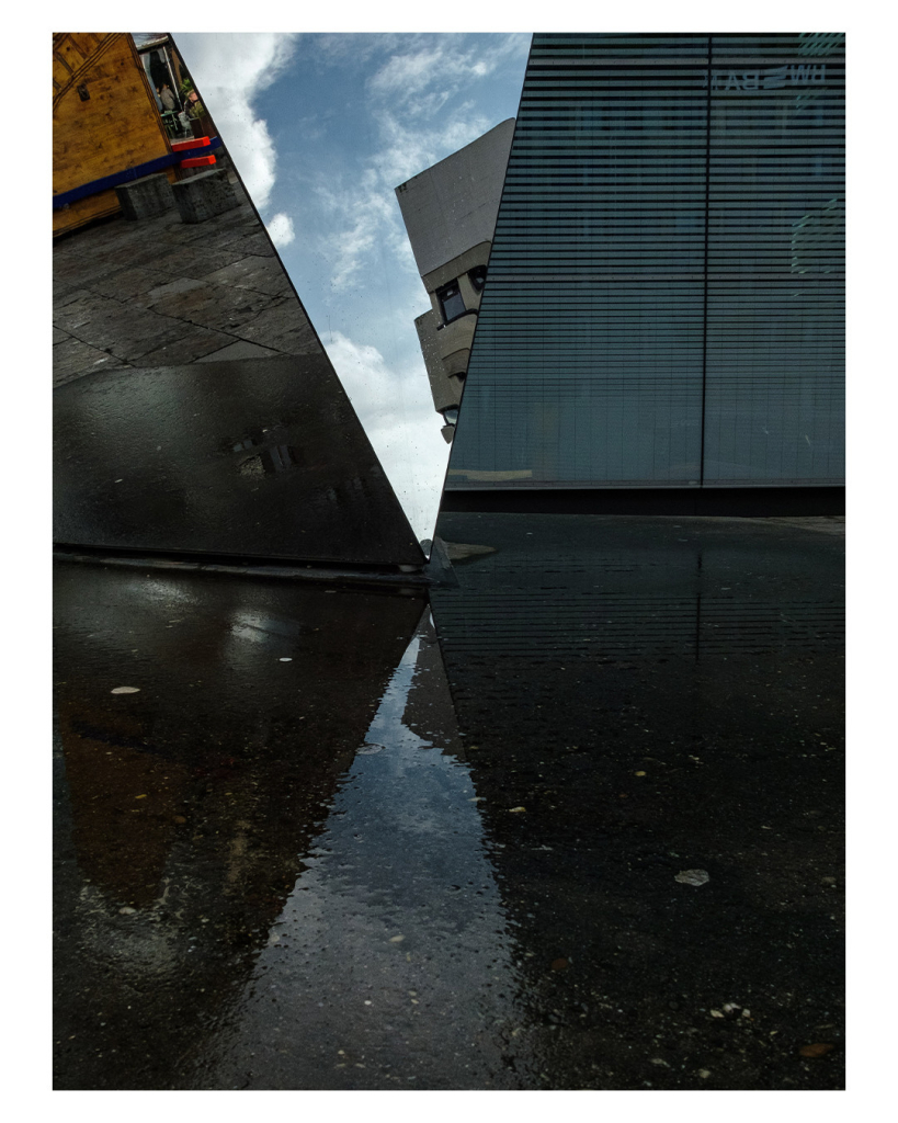 Foto im Hochformat. Eine dreidimensionale Skulptur in Pyramidenform. Die Spitze zeigt nach unten auf einen Sockel aus Beton. Die Seiten der Skulptur sind jeweils verspiegelt. Auf der Seite, die der Kamera zugewandt ist, spiegelt sich der Himmel und die obere Ecke eines Hauses. Unten auf dem Sockel spiegelt sich diese Ecke in einer Pfütze. 