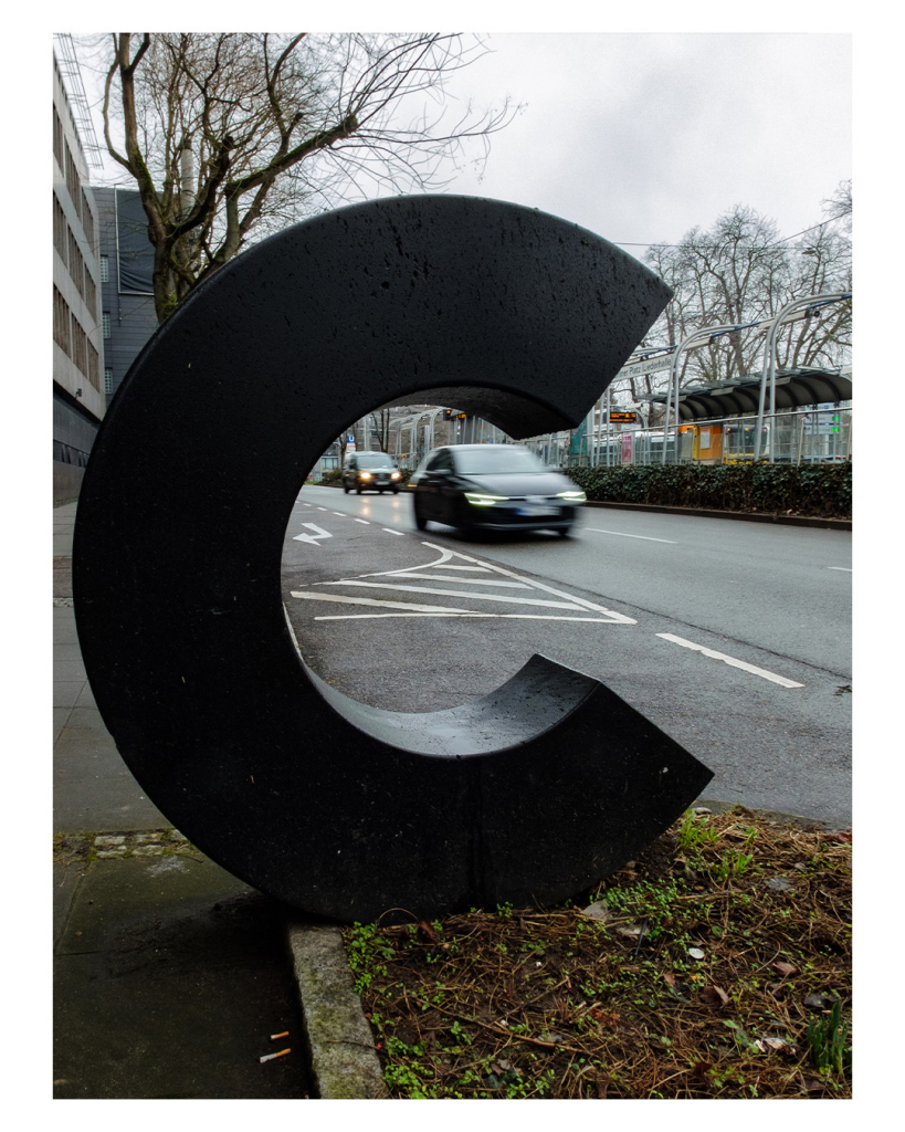 Foto im Hochformat. Eine Skulptur in Form des Buchstabens C steht an einer Hauptstraße in einer Stadt. Das C füllt fast das ganze Bild aus. Im C fährt gerade eine Auto auf der Straße, dieses ist durch lange Belichtungszeit unscharf. Der Himmel ist bedeckt. 