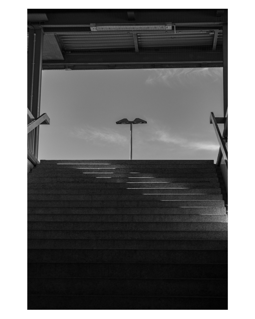 Schwarz-weiß Foto im Hochformat. Blick eine Treppe aus einer Unterführung hoch. Unten Treppe, links und rechts Pfosten, oben ein Dach, zusammen rahmen sie den Blick auf den fast wolkenlosen Himmel ein. Nur der obere Teil einer Straßenlaterne ragt ins Bild. Auf die Treppen fällt ein diagonaler Streifen Sonnenlicht. 