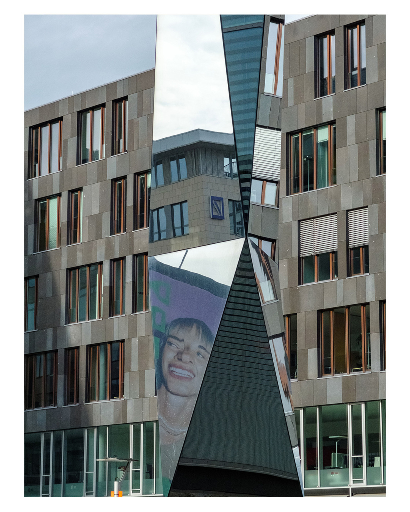 Foto im Hochformat. Ein bräunliches modernes Hochhaus. Davor steht eine Skulptur mit mehren Ebenen die alle verspiegelt sind. In diesen Ebenen sieht man teilweise andere Häuser und teilweise eine Person auf einer Werbetafel, die durch den Spiegel leicht verzerrt ist. 