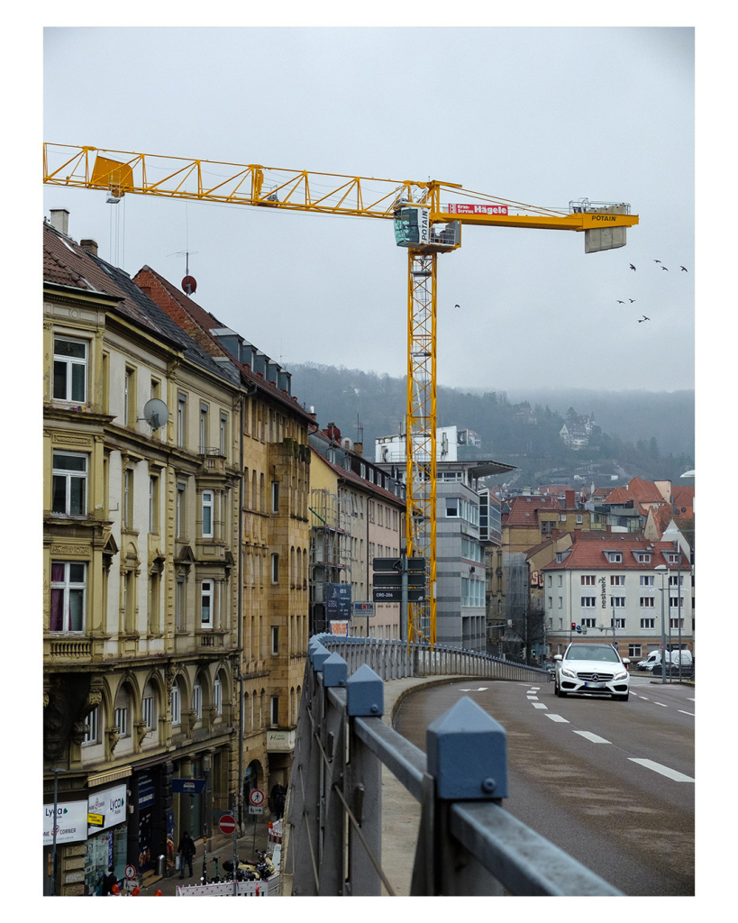 Foto im Hochformat. Blick entlang einer Straße in einer Großstadt aus der rechten unteren Ecke heraus verläuft das Geländer einer Brücke. Rechts davon verläuft die Straße mit wenigen Autos, links davon geht es in die Tiefe, unten verlauft eine Straße. An dieser stehen einige Altbauten. Ebenfalls unten steht ein gelber Kran, der über die Brücke und über die Altbauten ragt. Um den Kran fliegt ein Schwarm Vögel. Der Himmel ist komplett bewölkt und den hinten liegenden Berg in Nebel. 