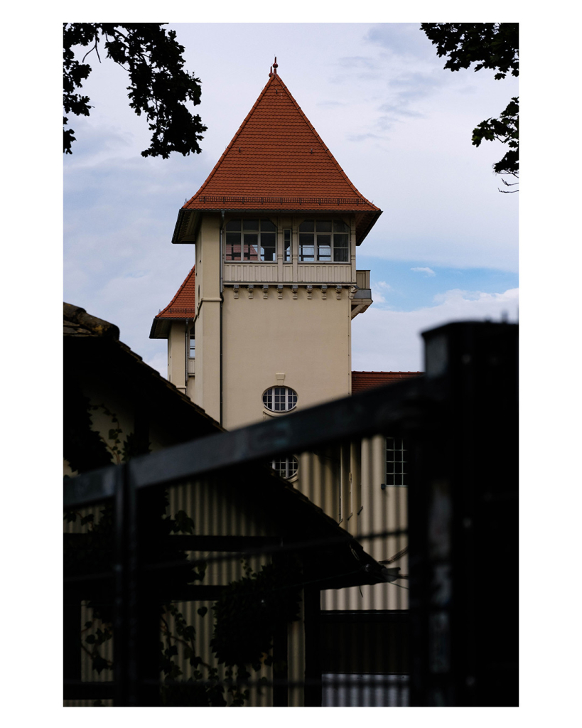 Foto im Hochformat. Ein eierfarbener Turm aus Stein mit rotem Spitzdach. Der Turm hat nur unter dem Dach Fenster und sieht deshalb aus wie ein Wachtturm. Oben ragen links und rechts Äste mit Blättern ins Bild. Unten verläuft ein hohes Gitter, welches dunkel ist, da es im Schatten liegt. Gitter und Äste rahmen den Turm ein. Der Himmel ist bewölkt. 