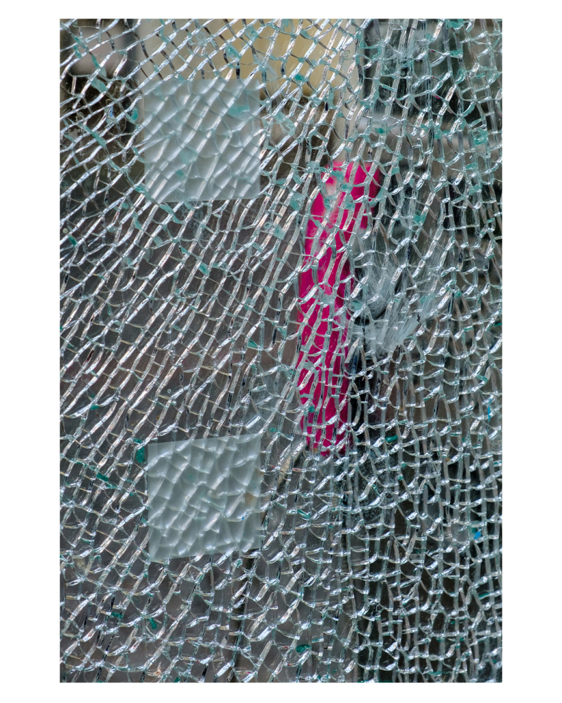 Foto im Hochformat. Nahaufnahme einer zerborstenen Glasscheibe. Sie ist übersät mit rissen. Hinter der Scheibe lässt sich der magenta farbene Telefonhörer einer Telefonzelle erahnen. 