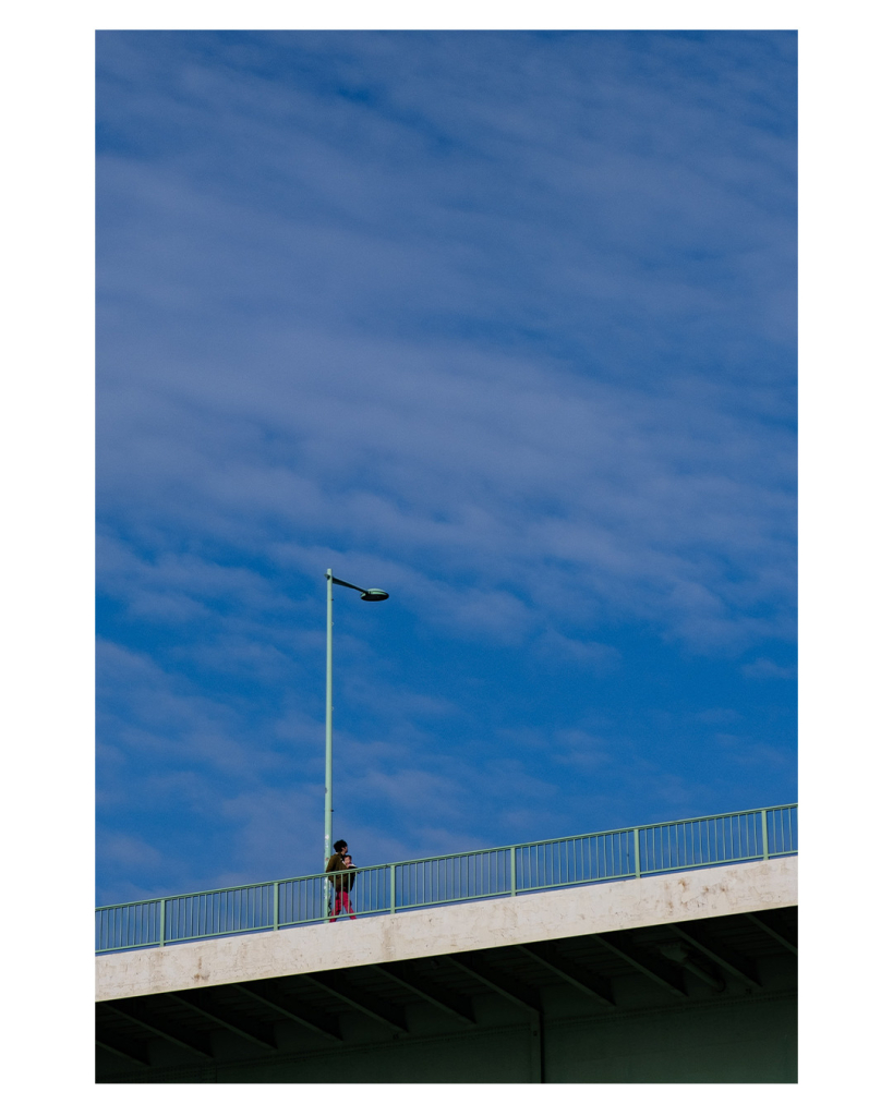 Foto im Hochformat. Teil einer Brücke von unten fotografiert. Eine einzelne Person läuft auf der Brücke und schaut in den Himmel. Dieser ist leicht bewölkt und nimmt den Großteil des Bildes ein. Hinter der Person ist eine einzelne grüne Straßenlaterne. 